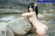 A woman in a bikini sitting on a rock by the water.