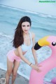 A woman in a bikini standing next to a pink flamingo on the beach.